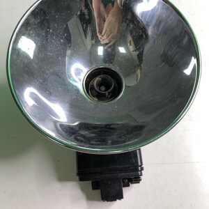 May include: A vintage camera flash unit featuring a large, circular, polished chrome reflector bowl with a central bulb socket. The base of the unit is constructed from black plastic and includes a mounting bracket for attachment to a camera body.