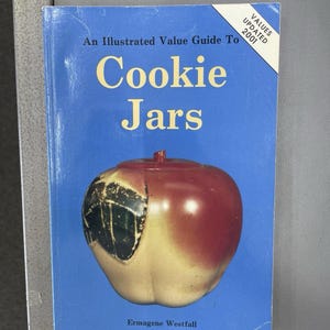May include: A softcover book titled An Illustrated Value Guide To Cookie Jars by Ermagene Westfall. The blue cover features a large red and yellow apple-shaped cookie jar with a black graphic detail and a white corner banner stating Values Updated 2001.