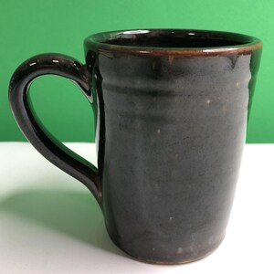 May include: A dark brown ceramic mug with a glossy finish and a smooth, cylindrical body. The mug features a curved handle on the left side and a slightly flared rim. The surface has subtle variations in color, showing hints of lighter brown tones.