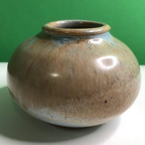 May include: A small, round ceramic vase with a smooth, glossy finish. The glaze features a blend of earthy tan and brown tones, with soft, muted blue accents around the rim and base. The vase has a wide, bulbous body and a narrow, circular opening.