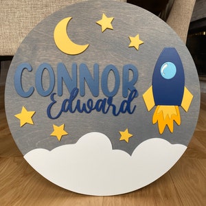 Puede incluir: Un letrero redondo de madera con fondo gris y una nube blanca en la parte inferior. El letrero presenta un cohete azul con llamas amarillas, una luna creciente amarilla y estrellas amarillas. El nombre "CONNOR Edward" est&aacute; escrito en letras azules.