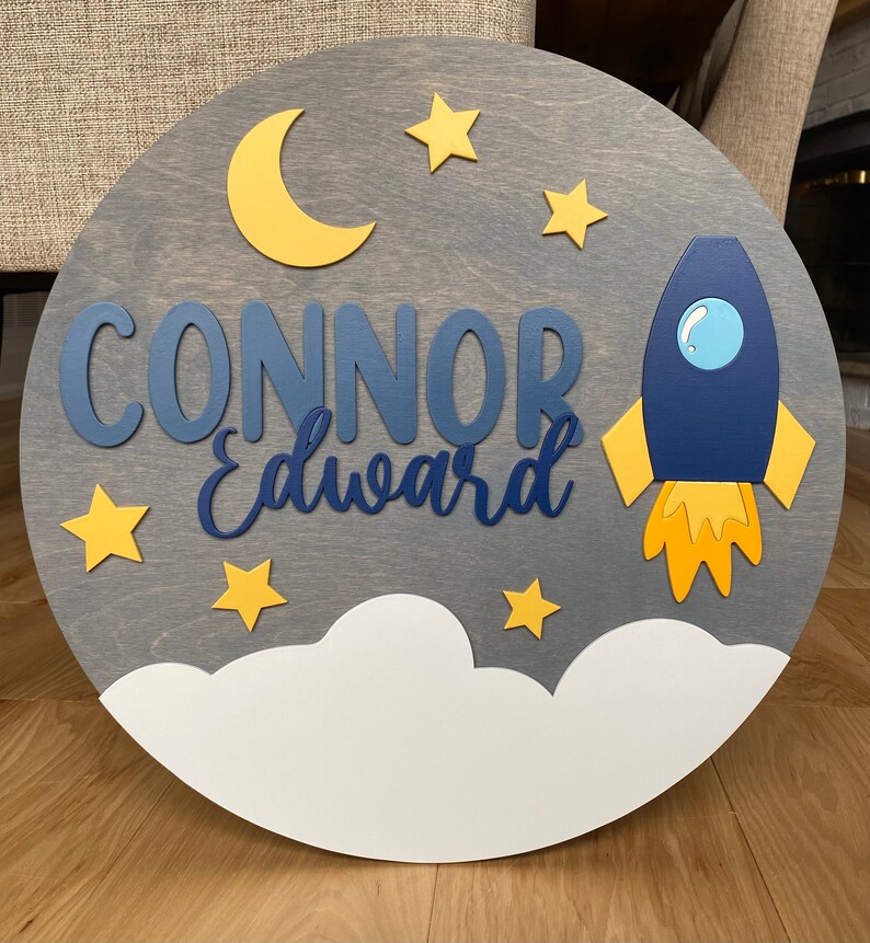Puede incluir: Un letrero redondo de madera con fondo gris y una nube blanca en la parte inferior. El letrero presenta un cohete azul con llamas amarillas, una luna creciente amarilla y estrellas amarillas. El nombre "CONNOR Edward" est&aacute; escrito en letras azules.