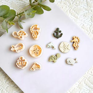 Op de afbeelding: Een verzameling koekjessnijders met zeethema's, waaronder een zeepaardje, walvis, dolfijn, anker en schildpad. De uitstekers zijn in beige, groen en bruin, gerangschikt op een wit oppervlak met een takje groen.