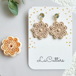 Op de afbeelding: Beige bloemen oorbellen met kristallen accenten, gepresenteerd op een witte kaart met gouden hartjes. Een bijpassende bloemvormige kleisnijder is ook aanwezig. De kaart heeft de tekst "Lü Cutters".