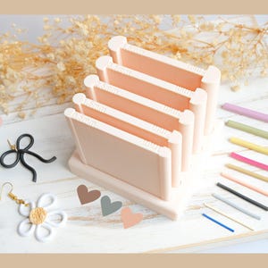 May include: A light peach-coloured craft tool with multiple slots labelled with measurements, alongside colourful rods and decorative items. The image includes a black bow, a white daisy earring, and heart-shaped cutouts.