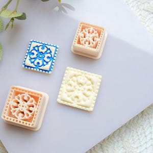 Può includere: Quattro timbri decorativi quadrati con disegni floreali. Un timbro è blu e bianco, due sono color pesca e uno è bianco sporco. I timbri sono su una superficie bianca e lilla chiaro.