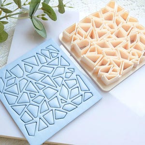 Puede incluir: Dos cortadores de galletas cuadrados. Uno es azul claro con un diseño geométrico, el otro es beige con un patrón geométrico en relieve similar. Los cortadores están sobre una superficie blanca con vegetación en el fondo.