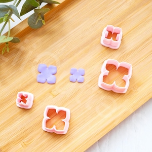Puede incluir: Un conjunto de cinco cortadores de galletas de plástico en forma de flor de color rosa y morado sobre una superficie de madera. Los cortadores son de diferentes tamaños y formas.