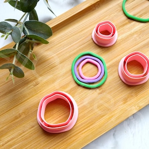 Puede incluir: Un juego de seis cortadores de galletas de plástico rosa y verde en varios tamaños y formas, incluyendo círculos, óvalos y flores. Los cortadores están dispuestos sobre una superficie de madera.