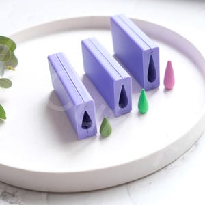 Può includere: Tre stampi in silicone viola per realizzare pezzi di resina a forma di lacrima. Gli stampi sono disposti in ordine di dimensione, con lo stampo più piccolo a destra e quello più grande a sinistra. Tre pezzi di resina a forma di lacrima sono mostrati accanto agli stampi, nello stesso ordine di dimensione.