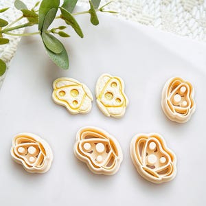 Può includere: Sei formine per biscotti beige e gialle a forma floreale sono disposte su una superficie bianca. Le formine hanno un aspetto a strati con un interno dettagliato. Un rametto di foglie verdi è nell'angolo in alto a sinistra.