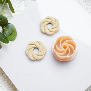 Puede incluir: Tres cortadores de galletas de color beige y melocotón sobre una superficie blanca. Dos son circulares con un diseño en espiral, y uno es un cortador en forma de flor. Hojas verdes en la esquina superior izquierda.