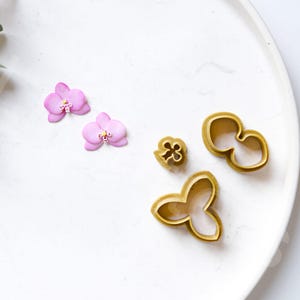 Puede incluir: Un juego de cuatro cortadores de galletas de color dorado con forma de flores de orquídea. Los cortadores están en un plato blanco con dos flores de orquídea rosas.