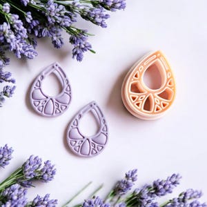 May include: Two pairs of purple and peach colored dangle earrings with a geometric design. The earrings are made of polymer clay and have a teardrop shape.