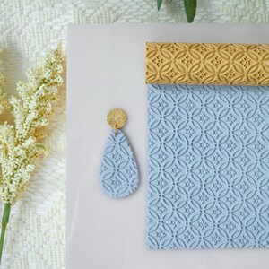 May include: A light blue teardrop-shaped earring with a gold glitter top, next to a matching textured blue sheet and a gold textured roller. The earring and sheet have a repeating pattern.