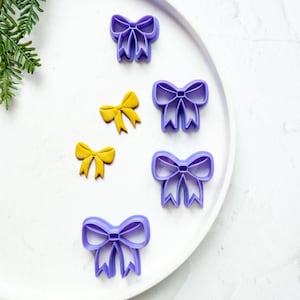 Puede incluir: Un juego de seis cortadores de galletas en forma de lazo, de color morado y amarillo, sobre un plato blanco. Los cortadores están dispuestos en semicírculo.