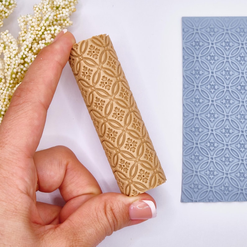 May include: A tan rolling pin with a repeating floral pattern is held in a hand. A blue rectangular mat with a similar pattern is next to the rolling pin. The rolling pin is cylindrical and has a detailed, embossed design.