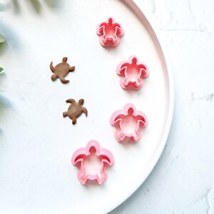 Puede incluir: Cuatro cortadores de galletas de tortuga de plástico rosa y dos cortadores de galletas de tortuga de plástico marrón en un plato blanco.