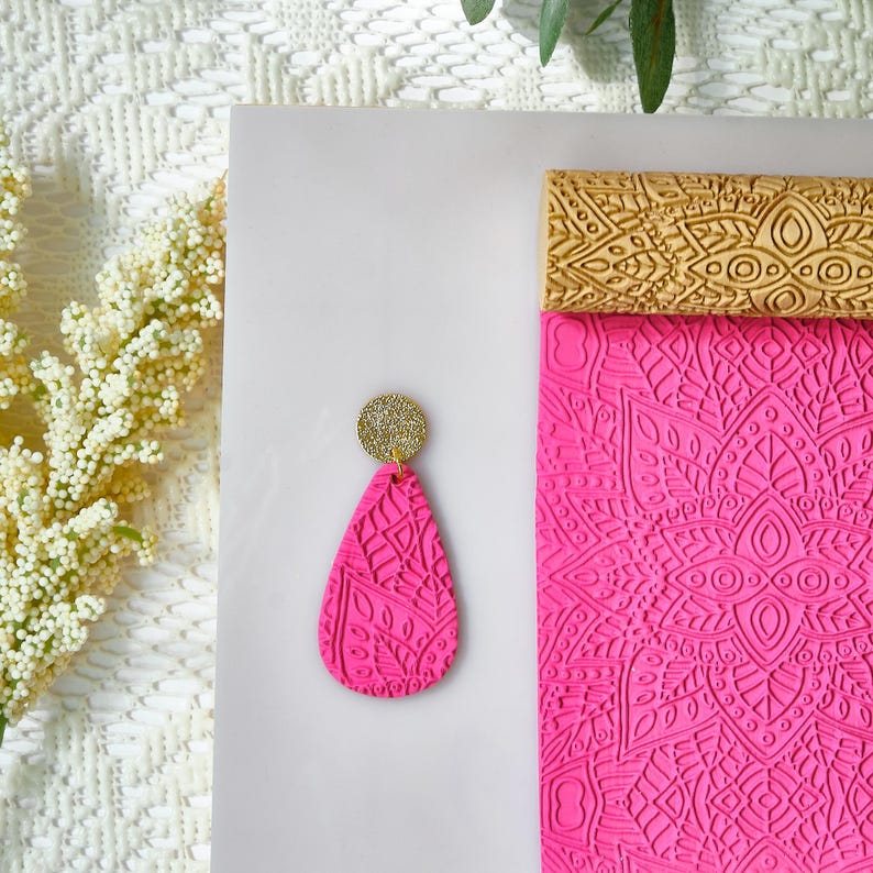 May include: A vibrant fuchsia teardrop earring with a gold glitter top. The earring features an embossed floral pattern. A matching fuchsia patterned block and a gold patterned block are also in the image.