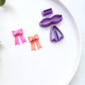Puede incluir: Cortadores de galletas de plástico morado en forma de lazo y cinta, con dos lazos rosas y dos lazos naranjas hechos con fondant.