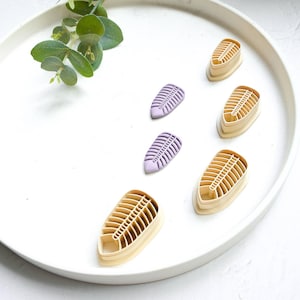 Può includere: Una collezione di formine per biscotti a forma di foglia in beige e lilla, disposte su un vassoio bianco con un rametto di verde. Le formine presentano un design a coste dettagliato, ideale per la cottura. Il tema è la cottura e l'artigianato.