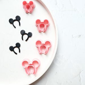 以下が含まれることがあります： 白い皿にピンクと黒のプラスチック製のミッキーマウスの耳のヘッドバンドが置かれています。ヘッドバンドは積み重ねられています。