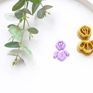 May include: Two gold-coloured metal flower cutters and a purple fondant flower on a white surface.