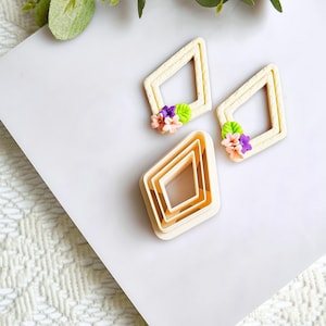 May include: A set of diamond-shaped polymer clay cutters with floral accents. The cutters are in various sizes, with three individual diamond frames and a stack of nested diamonds. The flowers are pink, purple, and white.