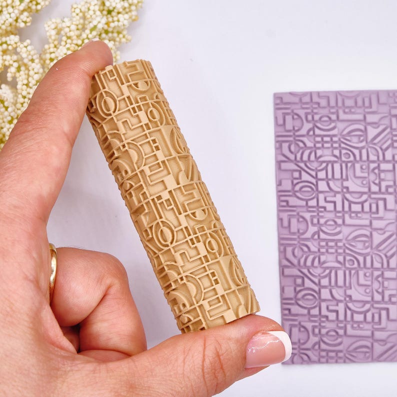 May include: A beige rolling pin with a geometric pattern is held in a hand. The pattern is also imprinted on a lavender rectangular surface. The rolling pin is cylindrical and features a repeating design of squares, circles, and lines. The design is raised, creating a textured effect.