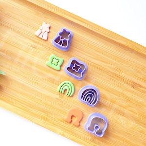 Könnte beinhalten: Ein Satz von acht violetten, grünen und orangefarbenen Plastik-Ausstechern auf einer Holzoberfläche. Die Ausstecher sind in Form eines Regenbogens, einer Blume, eines Kleeblatts, eines Hutes und anderer abstrakter Formen.