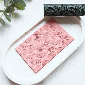 May include: A close-up of a pink and green rolling pin with a leaf pattern. The rolling pin is on a white surface.