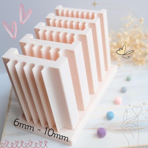 May include: A light pink nail art organizer with multiple slots, labeled from 6mm to 10mm. The organizer is designed to hold nail art supplies. The image includes decorative elements like hearts and a butterfly.
