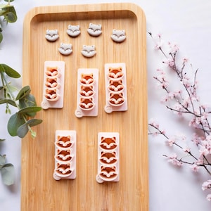 May include: A set of six silicone moulds for making fox-shaped cookies or fondant decorations. The moulds are pink and feature three fox faces stacked vertically in each mould. The moulds are arranged on a wooden surface with several individual fox-shaped moulds.