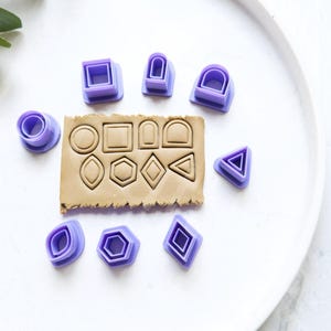 May include: A set of 12 purple plastic cookie cutters in various shapes, including circles, squares, diamonds, arches, and triangles. The cutters are arranged on a white surface with a brown clay base.