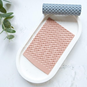May include: A light pink silicone rolling pin with a geometric pattern on a white ceramic tray. A blue silicone rolling pin with a similar pattern is in the background.
