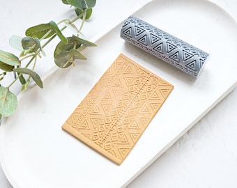 May include: A gray rolling pin and a flat piece of textured silicone, both with a geometric pattern of triangles and squares. The rolling pin and silicone are on a white surface.