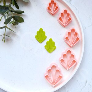 May include: A white plate with six pink and two green plastic cookie cutters shaped like seashells. The cookie cutters are arranged in a semi-circle on the plate.