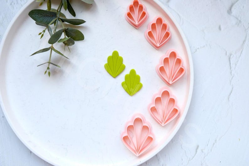 May include: A white plate with six pink and two green plastic cookie cutters shaped like seashells. The cookie cutters are arranged in a semi-circle on the plate.