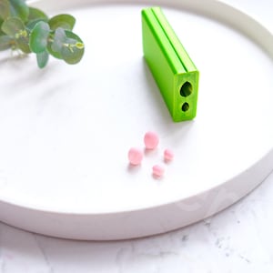 May include: A white ceramic plate with a green plastic rectangle with two holes in it. There are five pink round sweets on the plate.