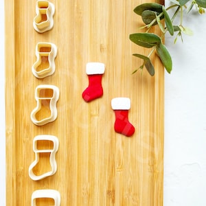 Puede incluir: Un juego de cinco cortadores de galletas de plástico blanco con forma de calcetines de Navidad y dos calcetines de Navidad de fondant rojo y blanco sobre una tabla de cortar de madera.