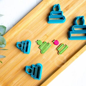 Puede incluir: Una bandeja de madera con cortadores de galletas con forma de libros y manzanas. Los cortadores son azules, verdes y rosas. La bandeja es de madera de color marrón claro. La bandeja mide unos 30 cm de largo.