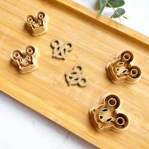 May include: Five cookie cutters on a wooden surface. The cutters are shaped like a bee, a mouse, and a bear. The cutters are made of a light brown plastic.