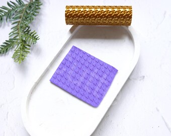 Può includere: Un tappetino in silicone testurizzato viola con un motivo ripetitivo di piccoli quadrati. Il tappetino è su una superficie bianca accanto a un rullo testurizzato dorato.