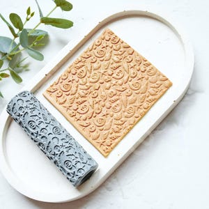 以下が含まれることがあります： グレーの模様入りローリングピンと、エンボス加工された花の模様が入った長方形のタン色の粘土片が、白い楕円形のトレイに置かれています。緑の葉の小枝が左上にあります。