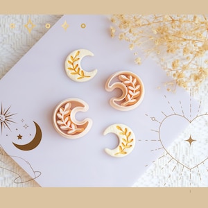 Pode incluir: Quatro cortadores de biscoitos em forma de meia-lua com desenhos botânicos. Os cortadores são de vários tamanhos e apresentam um exterior creme com um interior castanho claro. Os detalhes botânicos são dourados. O fundo inclui ilustrações celestiais.