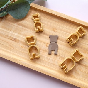 以下が含まれることがあります： クマの形をしたクッキーカッターが5つセットになっています。カッターは薄い茶色のプラスチック製で、木製の表面に並べてあります。カッターの1つは色が異なり、濃い茶色です。