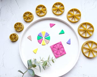 May include: A set of eight round cookie cutters in various sizes, all with a segmented design. The cutters are made of a light brown plastic and are arranged in a circle around a white plate. On the plate are several small pieces of colorful clay, including a pink, purple, green, and yellow circle, and several triangles in various colors. A square piece of pink clay is also on the plate, with a segmented design similar to the cookie cutters.