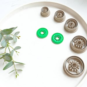May include: A set of seven cookie cutters on a white plate. The cutters are in the shape of a flower, a circle, and a kiwi. The kiwi cutters are green.