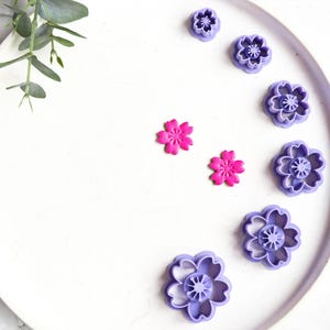 Puede incluir: Un juego de ocho cortadores de galletas con forma de flor de plástico morado y rosa en un plato blanco. Los cortadores de galletas están dispuestos en semicírculo.