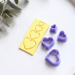 May include: A set of five heart-shaped cookie cutters. The cutters are made of purple plastic and come in various sizes. The largest cutter is on a yellow plastic base.